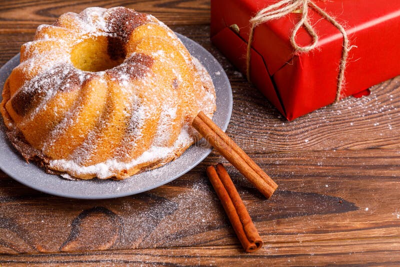 Traditional homemade cake decorated with cinnamon sticks and sugar powder. Gift box red. Christmas festive dessert on a brown wooden table. Traditional homemade cake decorated with cinnamon sticks and sugar powder. Gift box red. Christmas festive dessert on a brown wooden table.