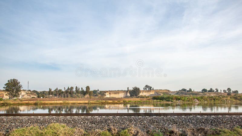 Train tracks beside a lake with a hill of land and trees with green foliage on a barren terrain in the background