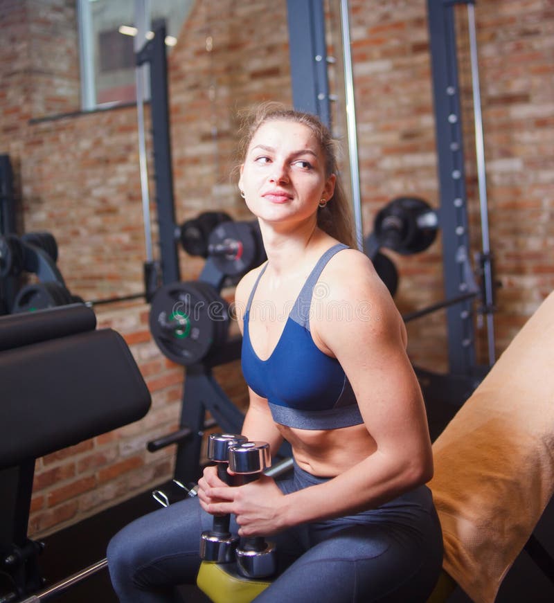 Training pause, sporty young woman in sportswear resting on bench holding dumbbells in her hands in the gym.