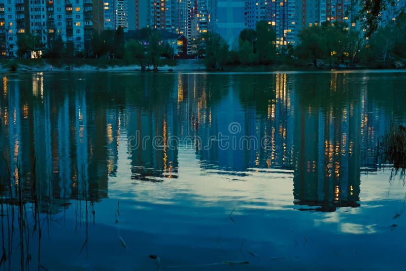 A tranquil evening scene of a lake reflecting city lights, buildings and water creating a mirrored effect at dusk. Reeds line the edges of the water.