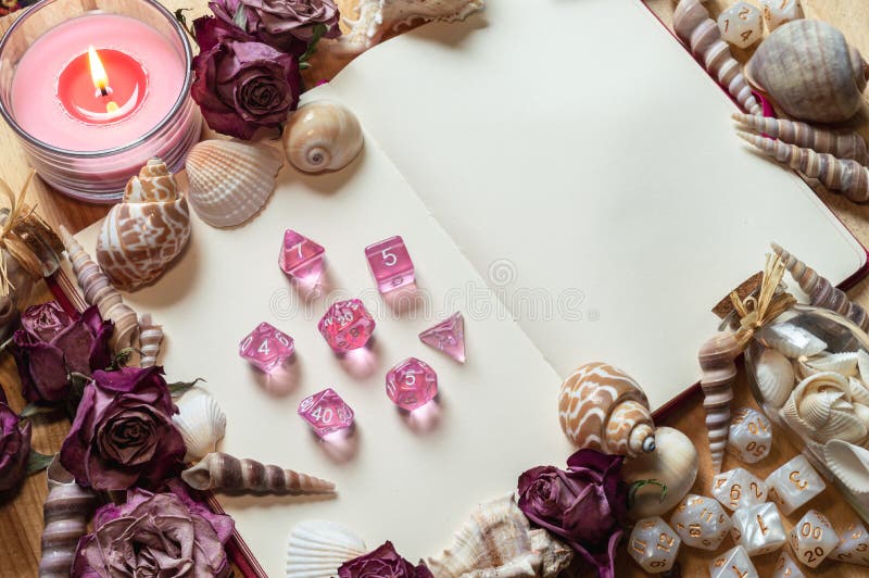 Transparent pink RPG gaming dice on an open sketchbook framed by seashells, dried pink roses, and white dice. Transparent pink RPG gaming dice on an open sketchbook framed by seashells, dried pink roses, and white dice