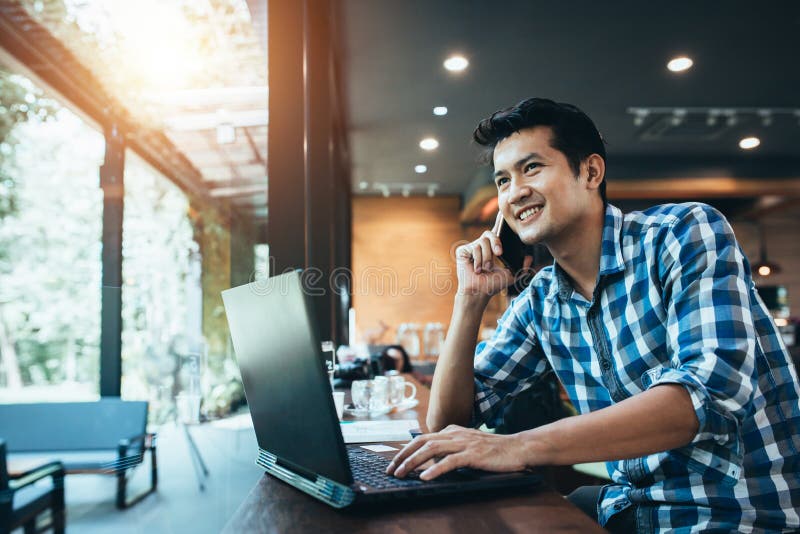 Travail indépendant d'homme asiatique sur le pavé tactile d'ordinateur tout en parlant au téléphone intelligent avec un sourire h
