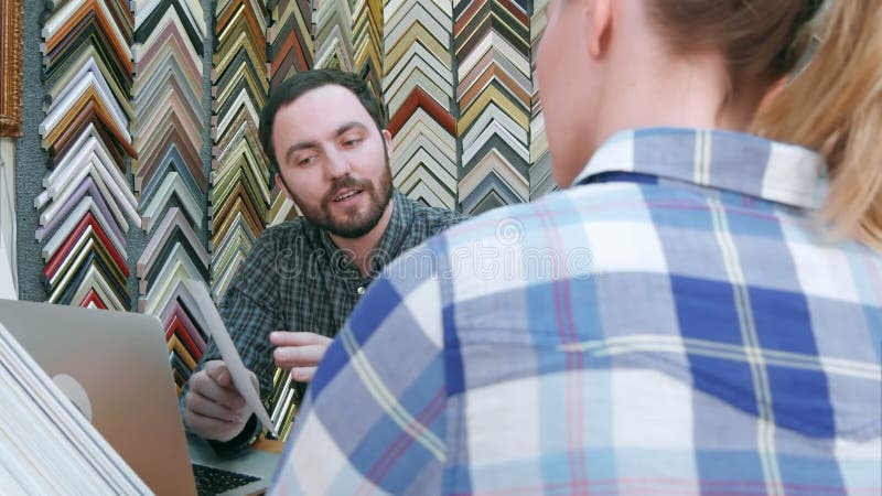Travailleur de jeune homme causant avec le client au sujet des détails de cadre photo, à l'aide de l'ordinateur portable sur le c banque de vidéos