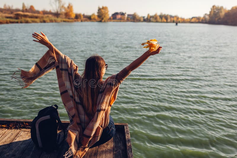 Traveler with backpack relaxing by autumn river at sunset. Young woman raised arms feeling free and happy stock photos Traveler with backpack relaxing by autumn river at sunset. Young woman raised arms feeling free and happy stock photos