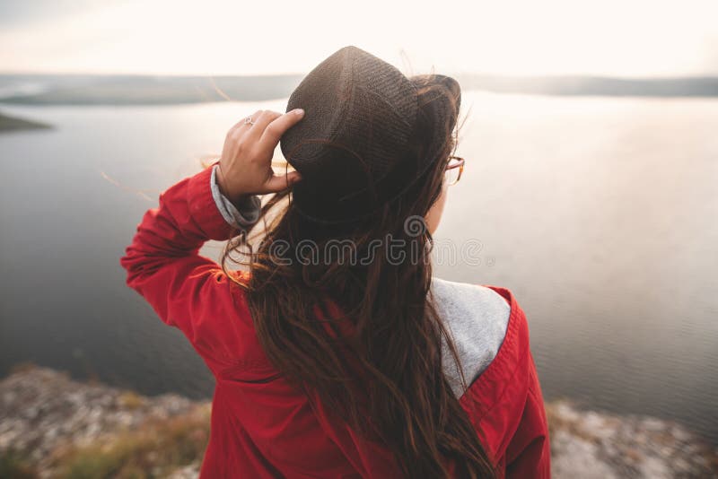 Traveler girl in hat and with windy hair standing in evening sunlight on top of rock mountain, enjoying beautiful sunset view on