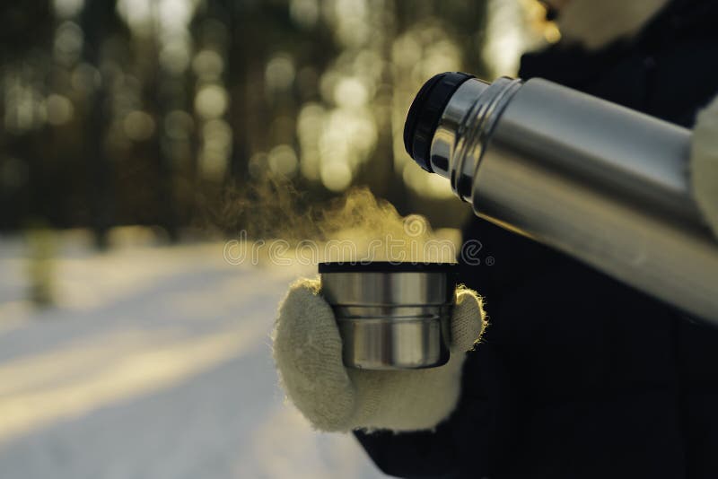 Traveler pours hot tea from a thermos into a mug, walking in snowy frozen winter forest at sunset
