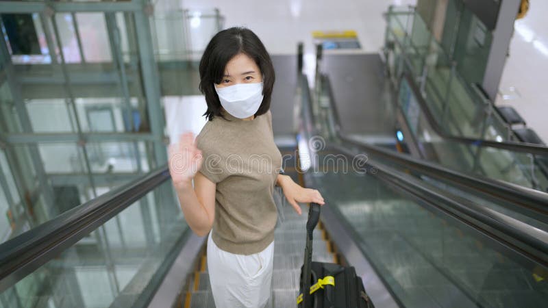 Traveler woman standing at escalator and turn back show hand waving gesture goodbye in the international airport terminal. Wearing