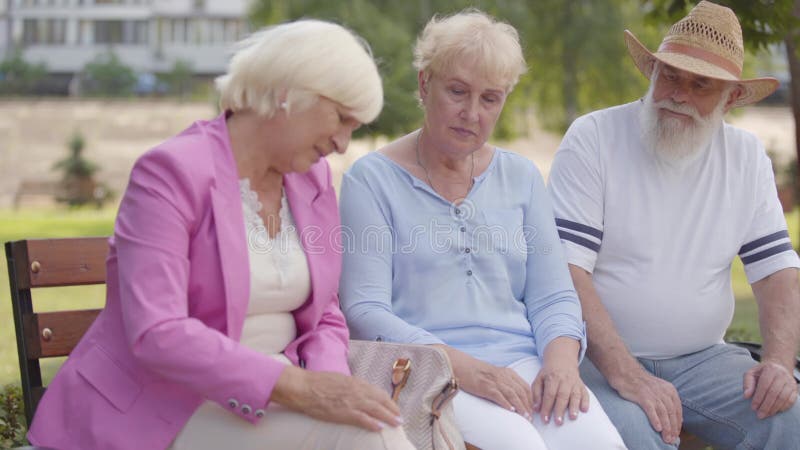 Tre mogna kaukasiska vänner som sitter i bänken i sommarparken Högre kvinna med blond hår som klagar på