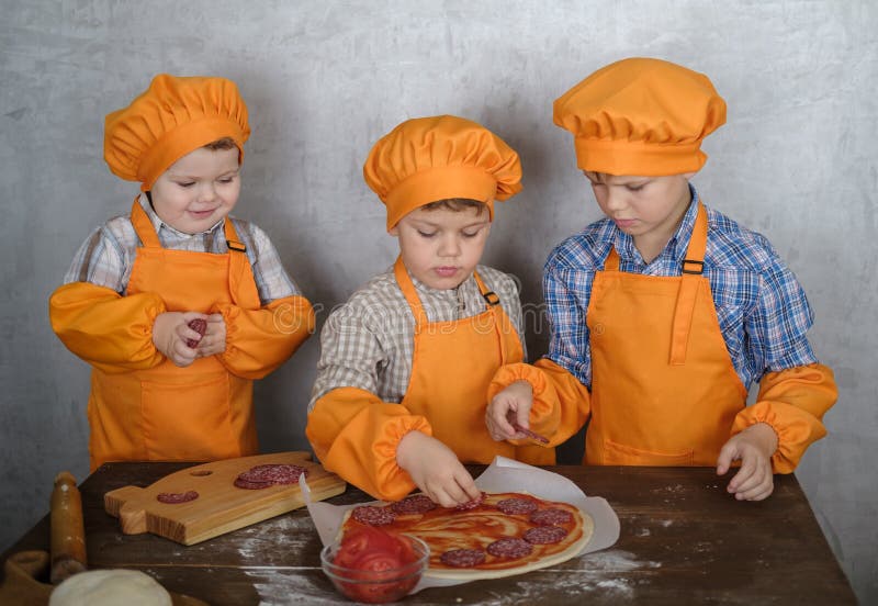 Tre ragazzi europei svegli vestiti come cuochi sono occupati cucinare la pizza i tre fratelli aiutano mia madre a cucinare la piz fotografia stock libera da diritti Tre ragazzi europei svegli vestiti come cuochi sono occupati cucinare la pizza i tre fratelli aiutano mia madre a cucinare la piz fotografia stock libera da diritti