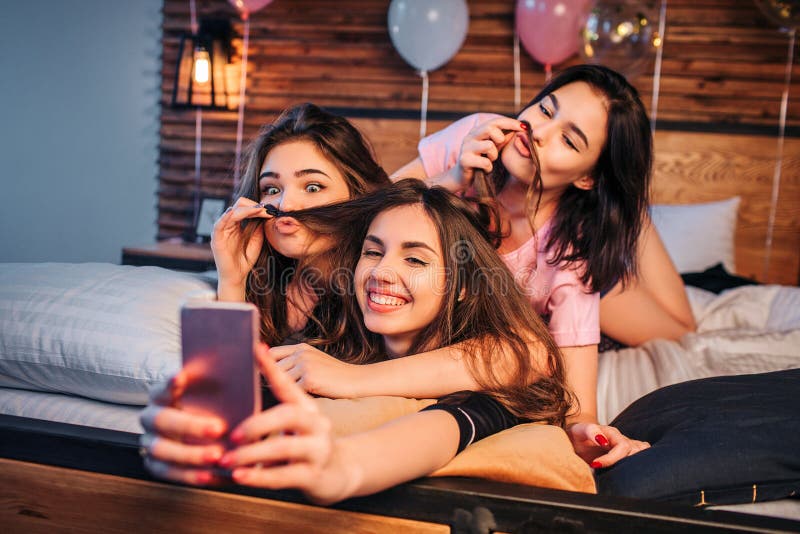 Tre skämtsamma unga kvinnor som tar selfie på säng i rum Lek för två modeller med hår av den tredje flickan De ser på kamera fotografering för bildbyråer Tre skämtsamma unga kvinnor som tar selfie på säng i rum Lek för två modeller med hår av den tredje flickan De ser på kamera fotografering för bildbyråer