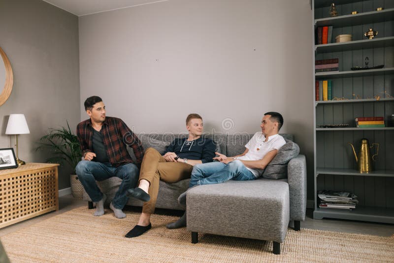 Tre trevliga unga killar sitter på soffan hemma och har roligt att prata arkivfoto Tre trevliga unga killar sitter på soffan hemma och har roligt att prata arkivfoto