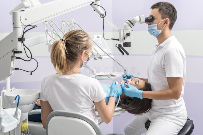 Patient in goggles on a dental chair and a dentist with assistant who sit next to her. Man looks on her teeth using a dental microscope and holds dental instruments. Woman holds an air water syringe. Patient in goggles on a dental chair and a dentist with assistant who sit next to her. Man looks on her teeth using a dental microscope and holds dental instruments. Woman holds an air water syringe.