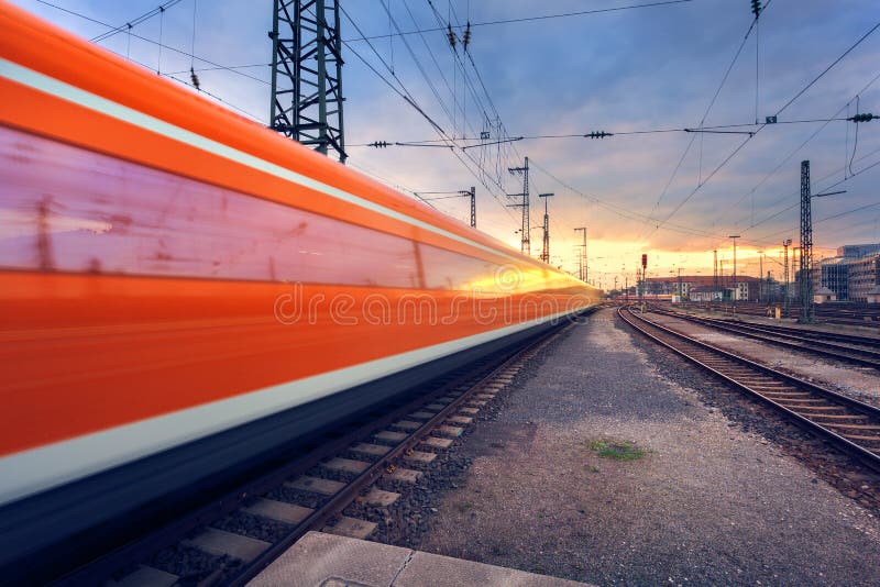 High speed red passenger train on railroad track in motion at sunset. Blurred commuter train. Railway station with cloudy sky. Railroad travel, railway tourism. Industrial landscape. Vintage toning. High speed red passenger train on railroad track in motion at sunset. Blurred commuter train. Railway station with cloudy sky. Railroad travel, railway tourism. Industrial landscape. Vintage toning