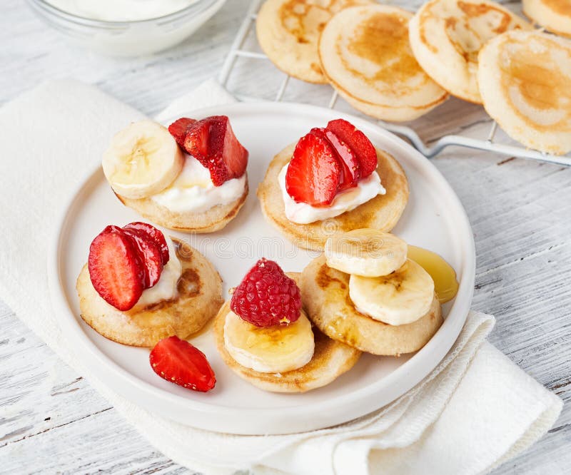 Trendy food pancake cereal with fruits, honey, yogurt. Heap of mini cereal pancakes in bowl on light background