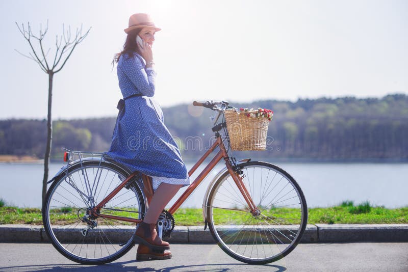 Trendy young woman stop to riding on her vintage bike with basket of flowers while focused chatting or talk on smart phone outside stock photo Trendy young woman stop to riding on her vintage bike with basket of flowers while focused chatting or talk on smart phone outside stock photo