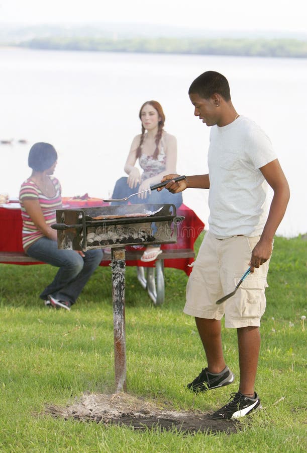 Teen boy tends the grill in the foreground, as two girls sit on a picnic table in the background. Vertical format. Teen boy tends the grill in the foreground, as two girls sit on a picnic table in the background. Vertical format.