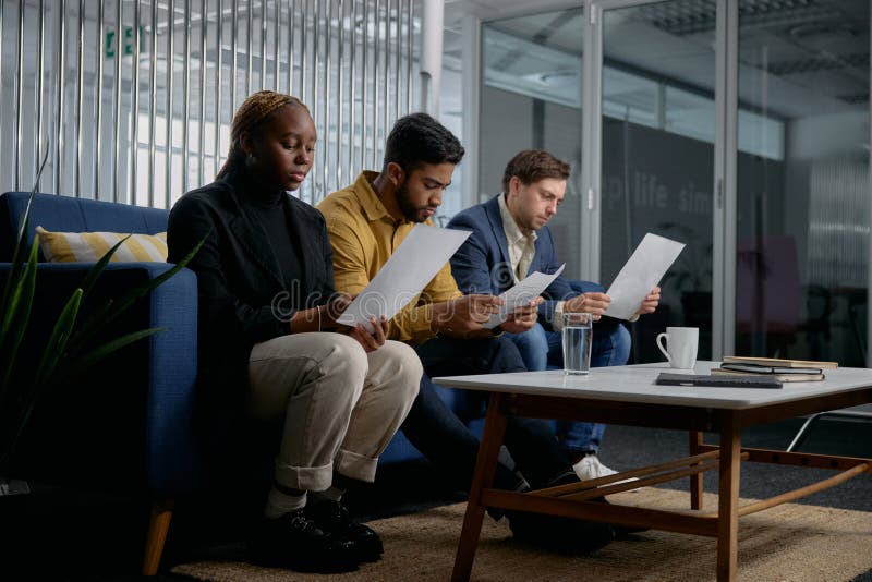 Three multiracial young adults wearing businesswear sitting on sofa and reading documents in office. Three multiracial young adults wearing businesswear sitting on sofa and reading documents in office