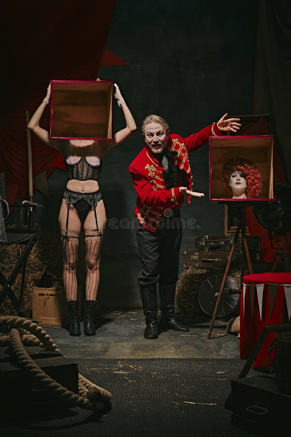 Tricks. Magician in red coat showing tricks with headless female assistant holding boxes over dark retro circus