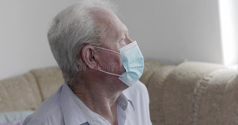 Trieste man in een medisch masker op sofa en door het raam kijkend