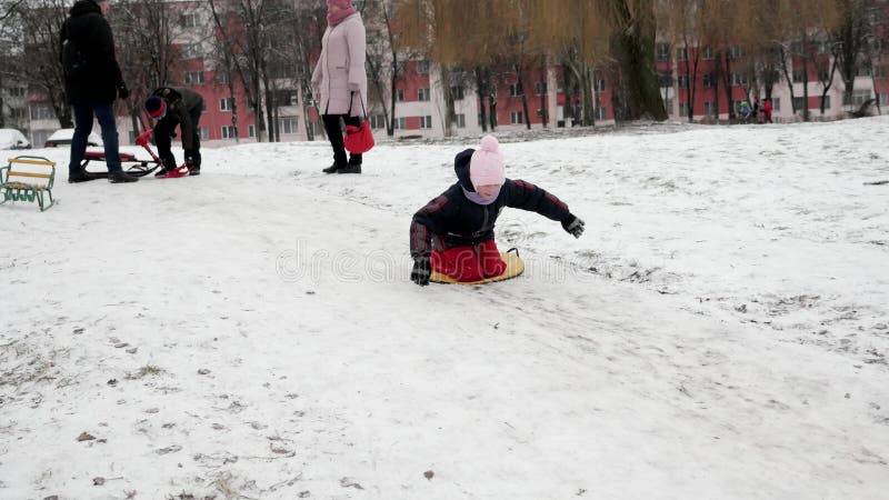 Trineo de chicas en una colina de nieve en el patio de un edificio de varios pisos. niños corriendo por la diapositiva : gomel bel