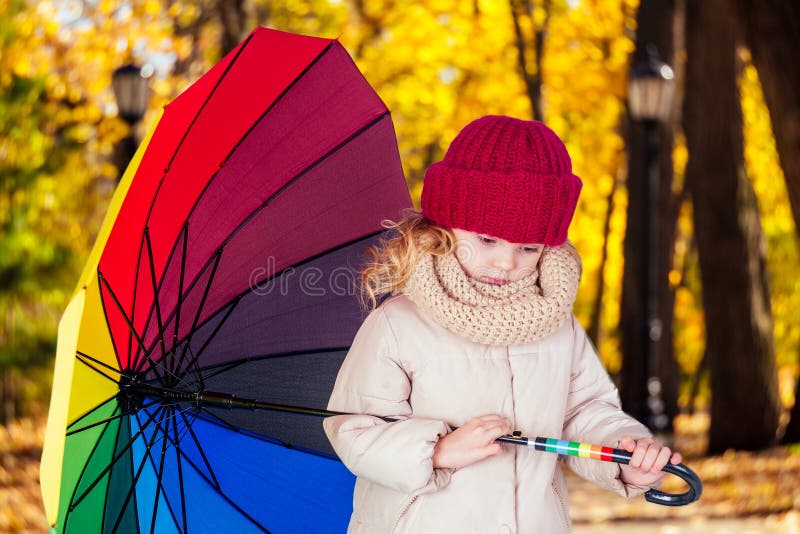 Triste blonde belle enfant avec parapluie coloré arc-en-ciel dans une veste grise dans le parc d'automne Jeune fille qui joue sur images libres de droits Triste blonde belle enfant avec parapluie coloré arc-en-ciel dans une veste grise dans le parc d'automne Jeune fille qui joue sur images libres de droits