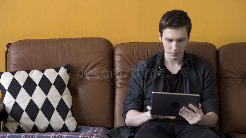 Trittbrettfahrer in schwarzem Hemd, der seine Tablette benutzt, während er zu Hause auf einem braunen Ledersofa sitzt Bestandsauf