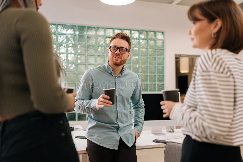 Three relaxed multiethnic coworkers standing in office, drinking coffee and talking cheerfully in workspace during break. Diverse male and female colleagues having conversation at modern open space. Three relaxed multiethnic coworkers standing in office, drinking coffee and talking cheerfully in workspace during break. Diverse male and female colleagues having conversation at modern open space.