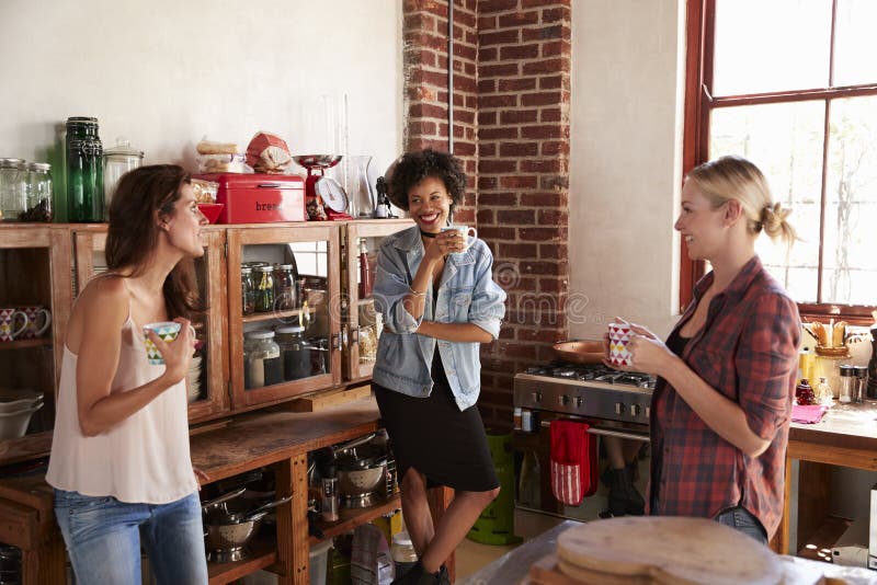 Trois jeunes amies adultes heureuses parlent la position dans la cuisine photographie stock libre de droits Trois jeunes amies adultes heureuses parlent la position dans la cuisine photographie stock libre de droits