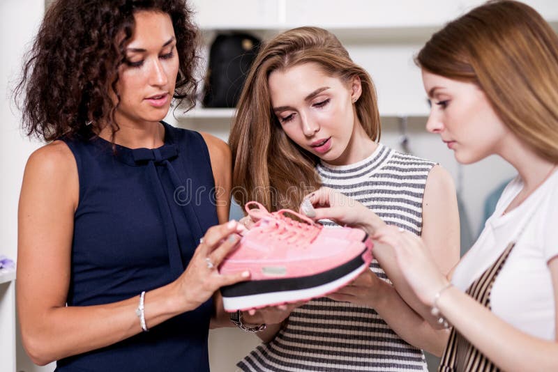 Trois jeunes amies examinant tenant de nouvelles paires de chaussures de sports se tenant dans la salle d'exposition de mode photos stock Trois jeunes amies examinant tenant de nouvelles paires de chaussures de sports se tenant dans la salle d'exposition de mode photos stock