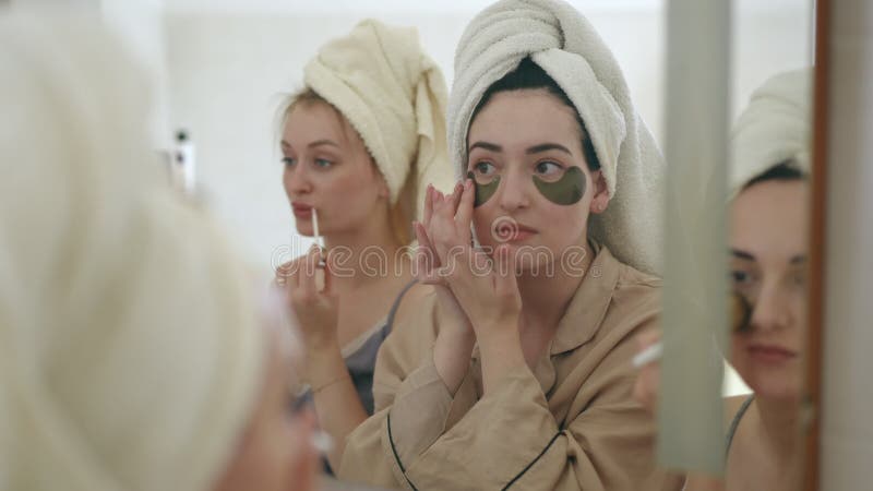 Trois jeunes femmes avec des serviettes sur la tête font des soins de beauté matinaux tout en se tenant devant un miroir dans