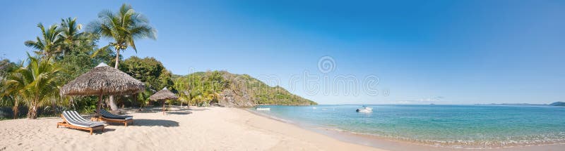 Tropical beach panorama with deckchairs, umbrellas, boats and palm tree