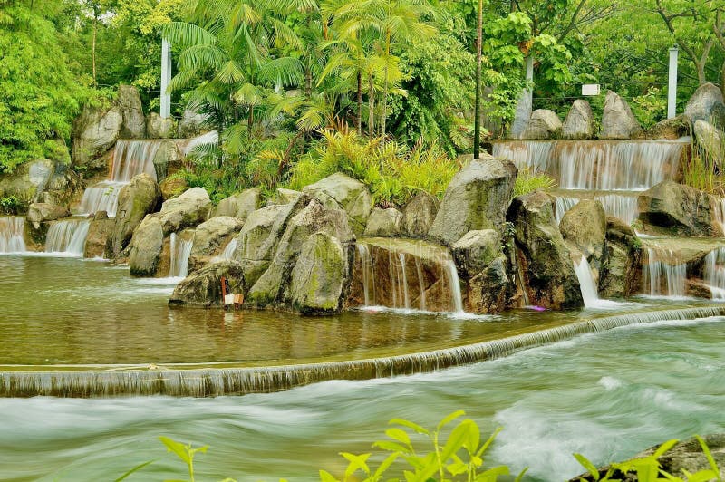 A vibrant and tranquil image capturing a beautifully designed multi-tiered man-made waterfall nestled within a lush, tropical park environment, likely in Singapore. The water cascades down moss-covered boulders and rocks, creating a soothing, layered effect that empties into a calm pool in the foreground. Beyond the falls, dense, verdant foliage, including palms and tropical plants, creates a refreshing backdrop. The lower part of the scene features a fast-moving stream or weir, hinting at the park's dynamic water features. This image perfectly encapsulates natural tranquility and the harmonious blend of nature and landscape architecture, making it ideal for themes of relaxation, travel, urban nature, and Southeast Asian landscapes. The long exposure effect on the moving water adds a dreamy, ethereal quality. Man made waterfall stock images, royalty-free photos and pictures