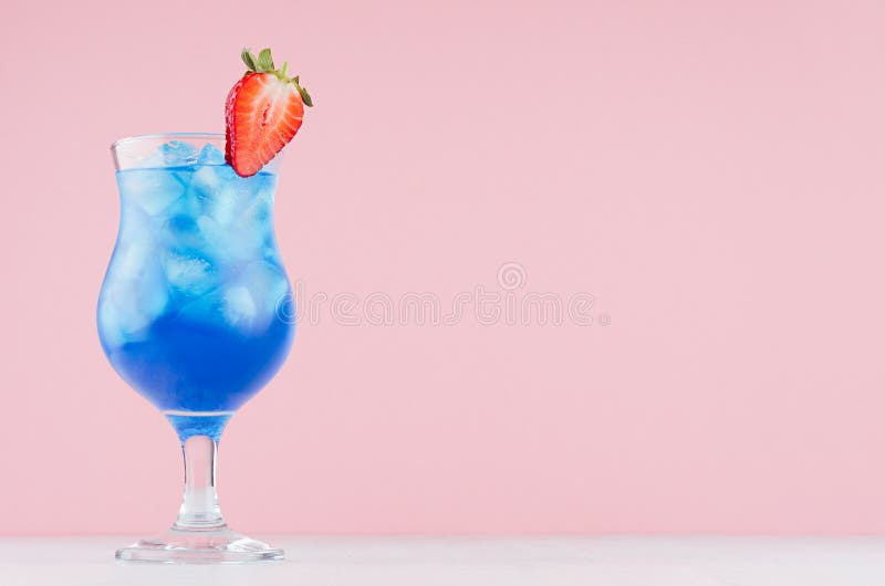 Tropical fresh blue drink with curacao, ice cubes, strawberry in exquisite misted wineglass on pastel pink wall, white table. stock image Tropical fresh blue drink with curacao, ice cubes, strawberry in exquisite misted wineglass on pastel pink wall, white table. stock image