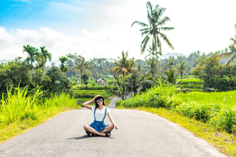 Tropisches Porträt der jungen glücklichen Frau mit Strohhut auf einer Straße mit Kokosnusspalmen und tropischen Bäumen Bali-Insel lizenzfreie stockfotografie Tropisches Porträt der jungen glücklichen Frau mit Strohhut auf einer Straße mit Kokosnusspalmen und tropischen Bäumen Bali-Insel lizenzfreie stockfotografie
