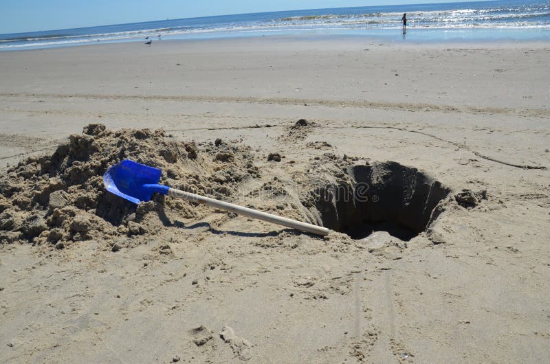A hole dug on the beach and left behind with a blue shovel and the ocean in the background. A hole dug on the beach and left behind with a blue shovel and the ocean in the background.