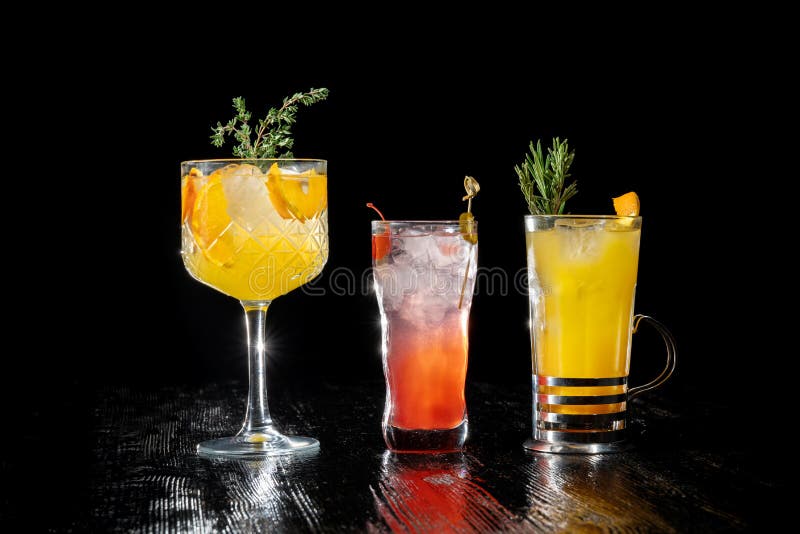 Three alcoholic cocktail with juice garnished. gin, apricot brandy and orange juice, garnished with rosemary, with orange slices. Back light. Glasses stand nearby. Three alcoholic cocktail with juice garnished. gin, apricot brandy and orange juice, garnished with rosemary, with orange slices. Back light. Glasses stand nearby.