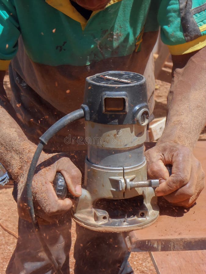 Metal cased, black and silver heavy-duty electric Woodcarving Router in use; solid grip on side handles as it carves shape in wood propelling airborne dust, some landing on hands. Metal cased, black and silver heavy-duty electric Woodcarving Router in use; solid grip on side handles as it carves shape in wood propelling airborne dust, some landing on hands.