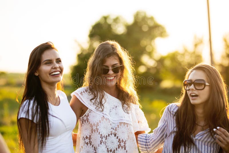 Três jovens mulheres de cabelo escuro bonitas nos óculos de sol vestidos na roupa bonita estão sorrindo e estão andando exteriore imagem de stock royalty free Três jovens mulheres de cabelo escuro bonitas nos óculos de sol vestidos na roupa bonita estão sorrindo e estão andando exteriore imagem de stock royalty free