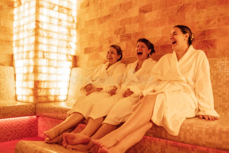 Three young women in white bath robes are sitting in a salt room. Attractive female friends laugh and relax during a spa treatment. Clients of a beauty salon or clinic at a halotherapy session. Three young women in white bath robes are sitting in a salt room. Attractive female friends laugh and relax during a spa treatment. Clients of a beauty salon or clinic at a halotherapy session