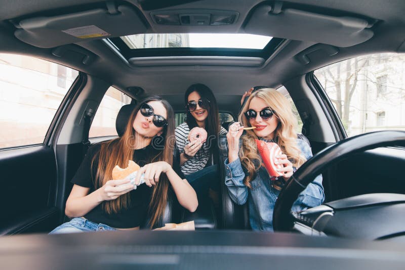 Três meninas que conduzem em um carro e que têm o divertimento que come o fast food na viagem por estrada do curso imagem de stock Três meninas que conduzem em um carro e que têm o divertimento que come o fast food na viagem por estrada do curso imagem de stock