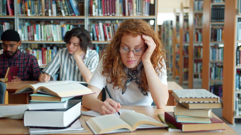 Trött rödhårig flicka som studerar i biblioteket och sedan sover på skrivbordet och frigör inomhus arkivfilmer