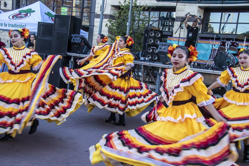 2021_10_09 Tulsa USA Dancing Hispanic girls swirl their beautiful yellow costumes as they dance at a street Festival in front of a stange and speakers with a mother watchin.g them and man videoing them-motion blur. 2021_10_09 Tulsa USA Dancing Hispanic girls swirl their beautiful yellow costumes as they dance at a street Festival in front of a stange and speakers with a mother watchin.g them and man videoing them-motion blur