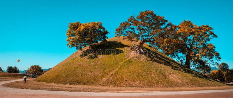 Tumuli royal tomb in autumn foliage backgroung. Gyeongju, South Korea.The Gyeongju Historic Areas of South Korea were designated as a World Heritage Site by UNESCO in 2000, ref 976. Teal and orange view.