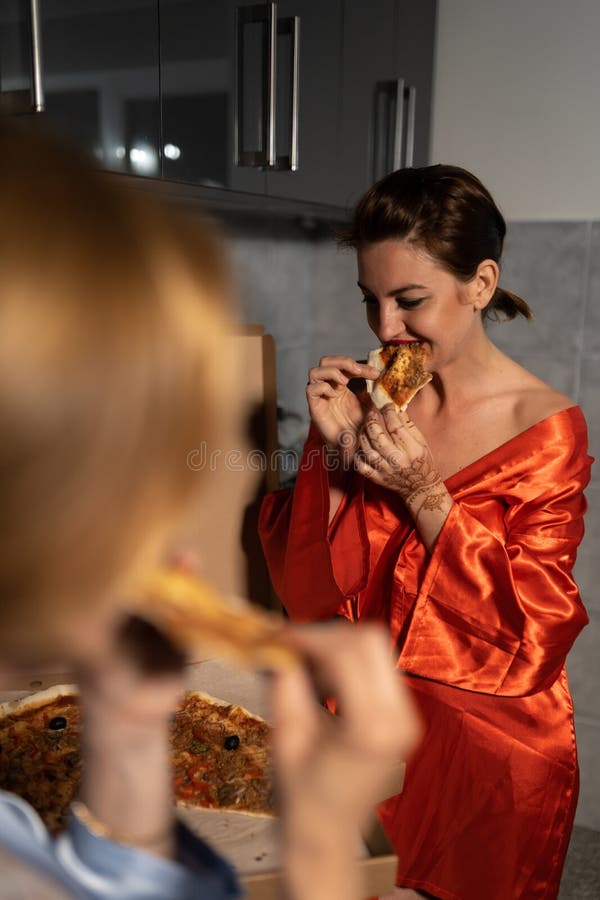 Tv? flickv?nkvinnor som ?ter pizza och tycker om en pratstund f?r aftonparti, innan att g? ut - en b?rande bl? morgonkappa arkivbilder Tv? flickv?nkvinnor som ?ter pizza och tycker om en pratstund f?r aftonparti, innan att g? ut - en b?rande bl? morgonkappa arkivbilder