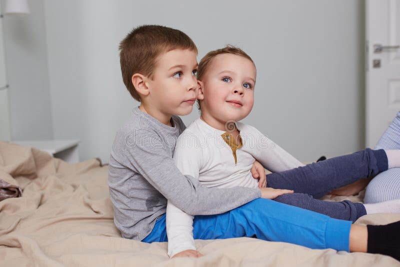 Tv? lilla br?der sitter huging p? s?ngen med den beigea filten i det ljusa hemtrevliga sovrummet arkivfoton Tv? lilla br?der sitter huging p? s?ngen med den beigea filten i det ljusa hemtrevliga sovrummet arkivfoton