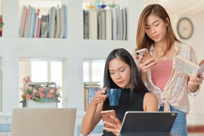 Två asiatiska kvinnliga studenter håller en kaffekopp och tittar på en smartphone för att förbereda sig för akademiska studier arkivbilder