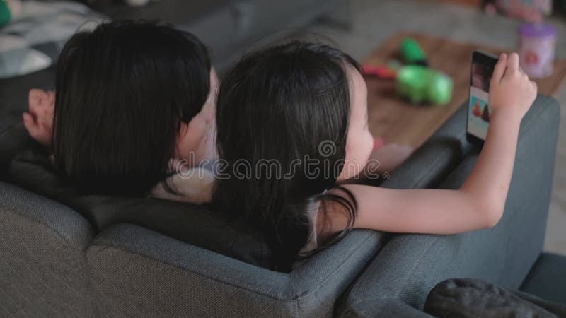 Två asiatiska, thai, svarta hårflickor som är syskon och sitter på en grå soffa i husets vardagsrum och båda tittar på