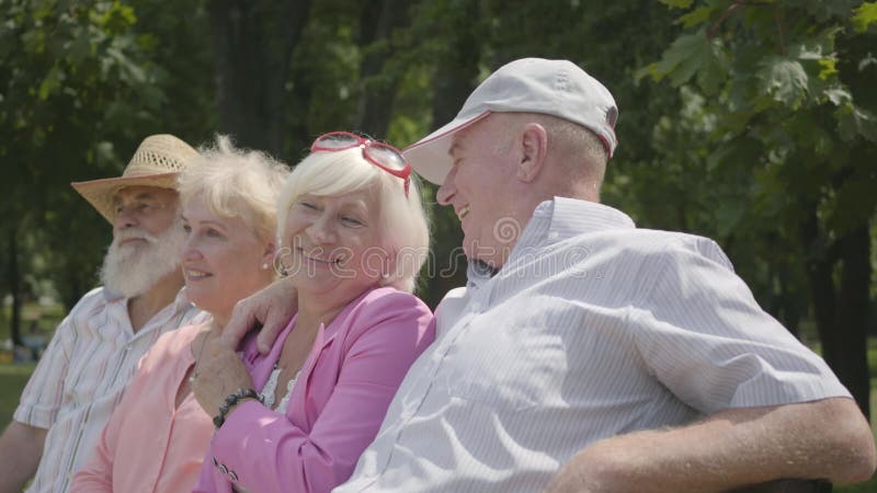 Två förtjusande mogna par som talar och ler sammanträde på bänken i sommaren, parkerar Dubbelträff av höga par