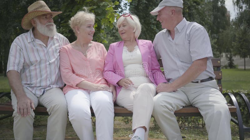 Två gulliga mogna par som talar och ler sammanträde på bänken i sommaren, parkerar Dubbelträff av höga par