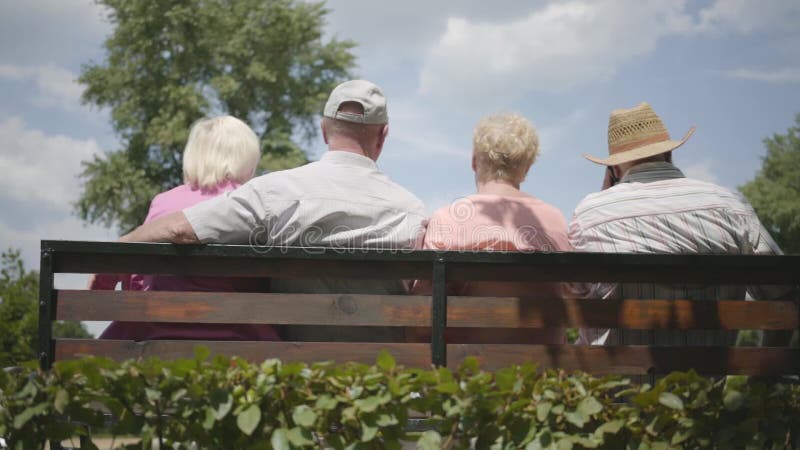 Två gulliga vuxna par som sitter på bänken och tillsammans talar Moget folk som vilar det fria gladlynt pension?r